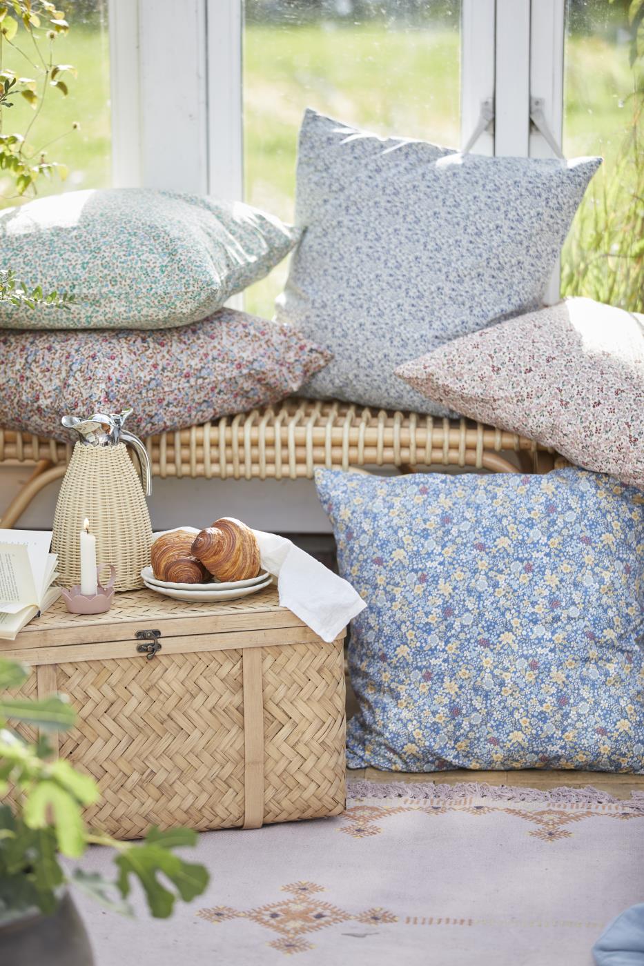 Naturfärgad termoskanna i polyrattan på ett litet bord i ett ljust uterum med kuddar och flätade möbler.