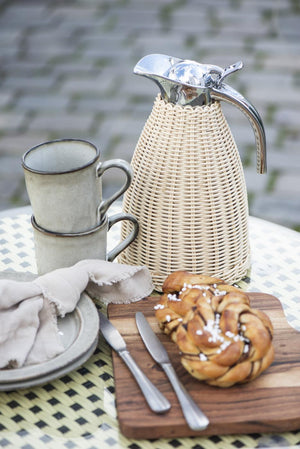 Flätad termoskanna från Ib Laursen på utomhuskaffebord med keramikmuggar, en croissant och rustikt servis.