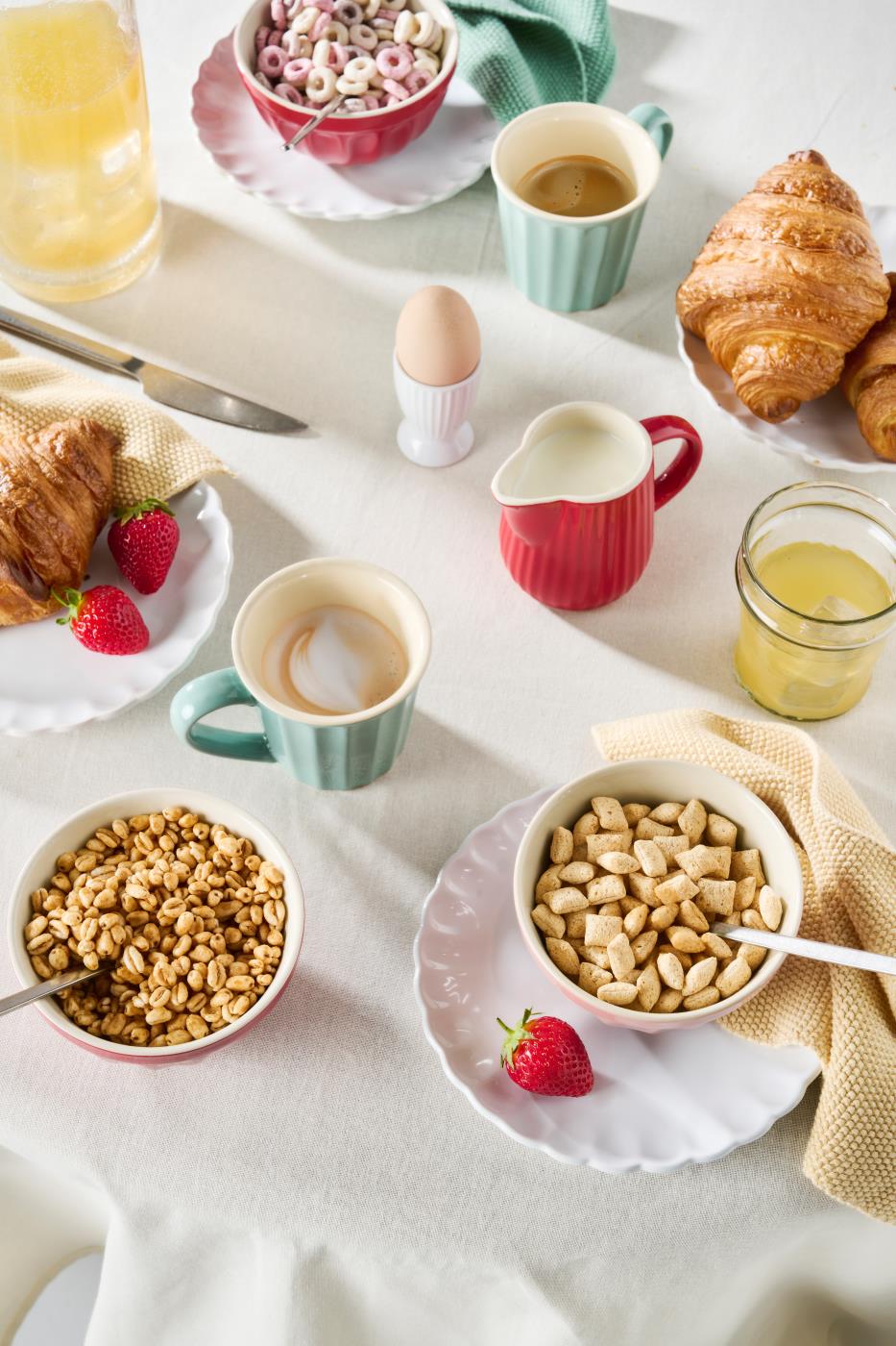 Mjölkkanna i jordgubbsrött och myntagrönt på frukostbordet med croissanter, müsli, kaffe och färsk frukt