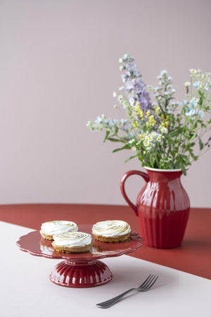 Röd myntakanna med blommor på sommarbord med kakfat och rosa bakgrund