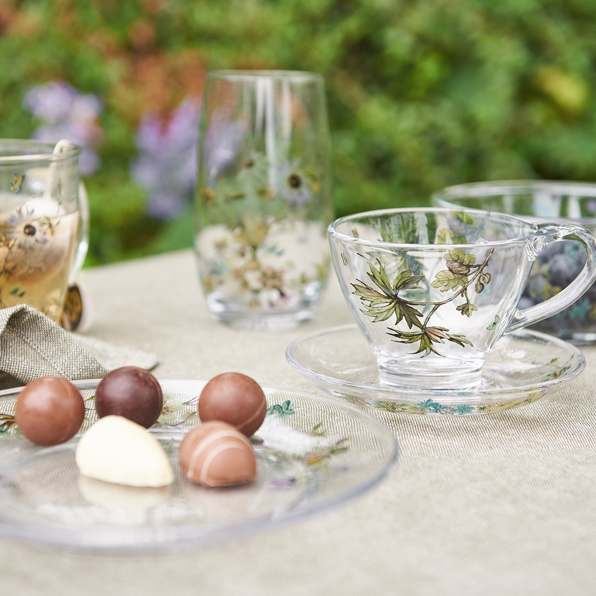 Blommig glaskopp med fat från Speedtsberg på ett somrigt kaffebord med matchande glas och små chokladbitar.