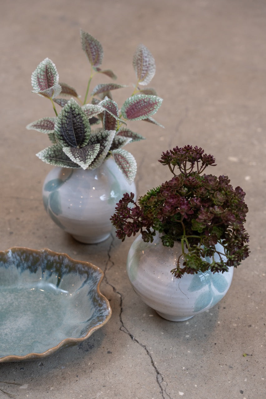 Konstgjord kvist med fläckiga gröna och rosa blad i en liten keramikvas tillsammans med mörka höstblommor.