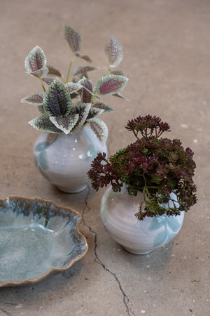 Konstgjord kvist med fläckiga gröna och rosa blad i en liten keramikvas tillsammans med mörka höstblommor.