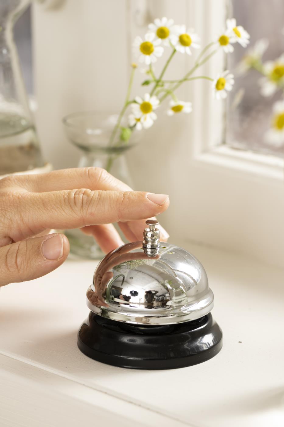 Hand som trycker på en blank diskklocka från Ib Laursen på en ljus fönsterbänk med blommor