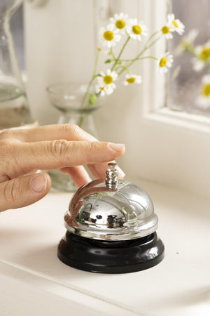 Hand som trycker på en blank diskklocka från Ib Laursen på en ljus fönsterbänk med blommor