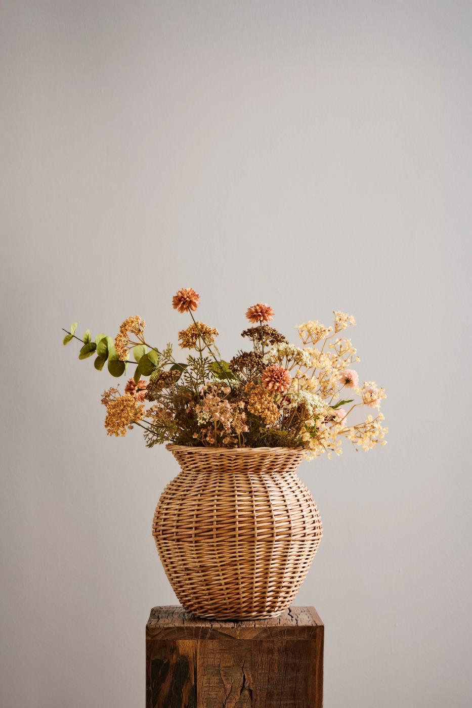 Höstbukett med konstgjord eukalyptus och blommor i gyllene, bruna och krämfärgade nyanser i en flätad vas.